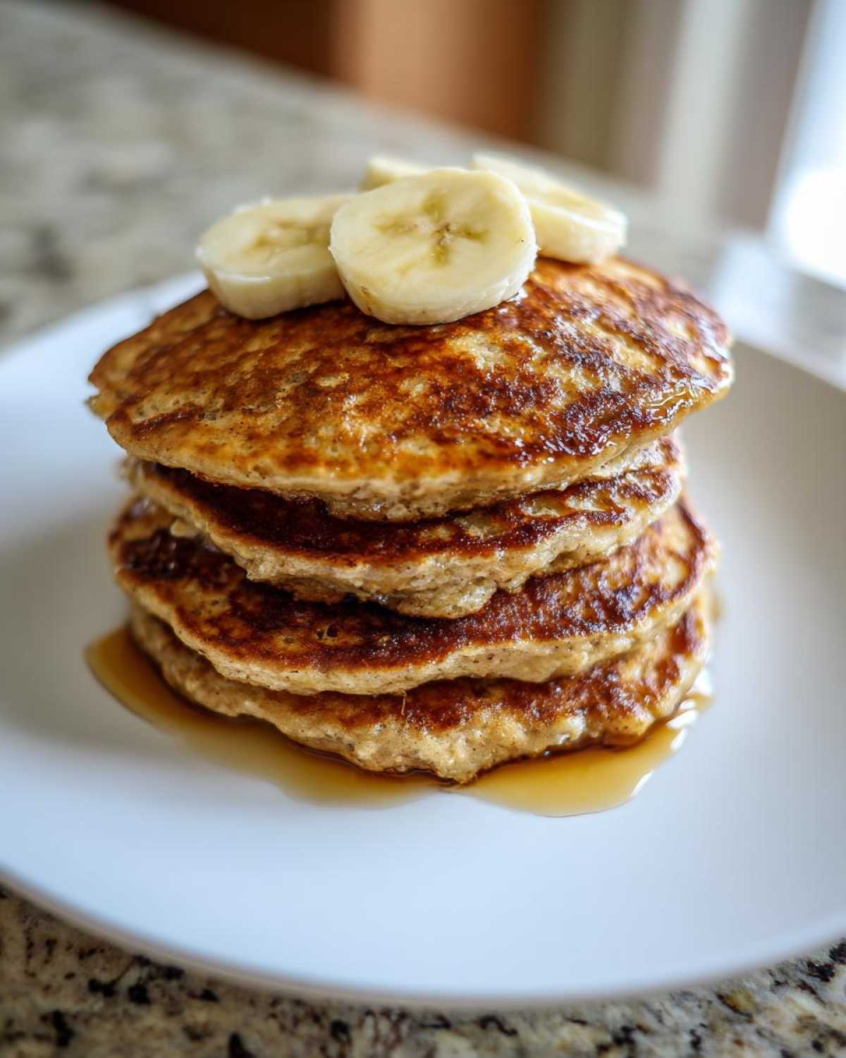 A tall stack of golden-brown Vegan Banana Oat Pancakes drizzled with syrup and topped with fresh banana slices.