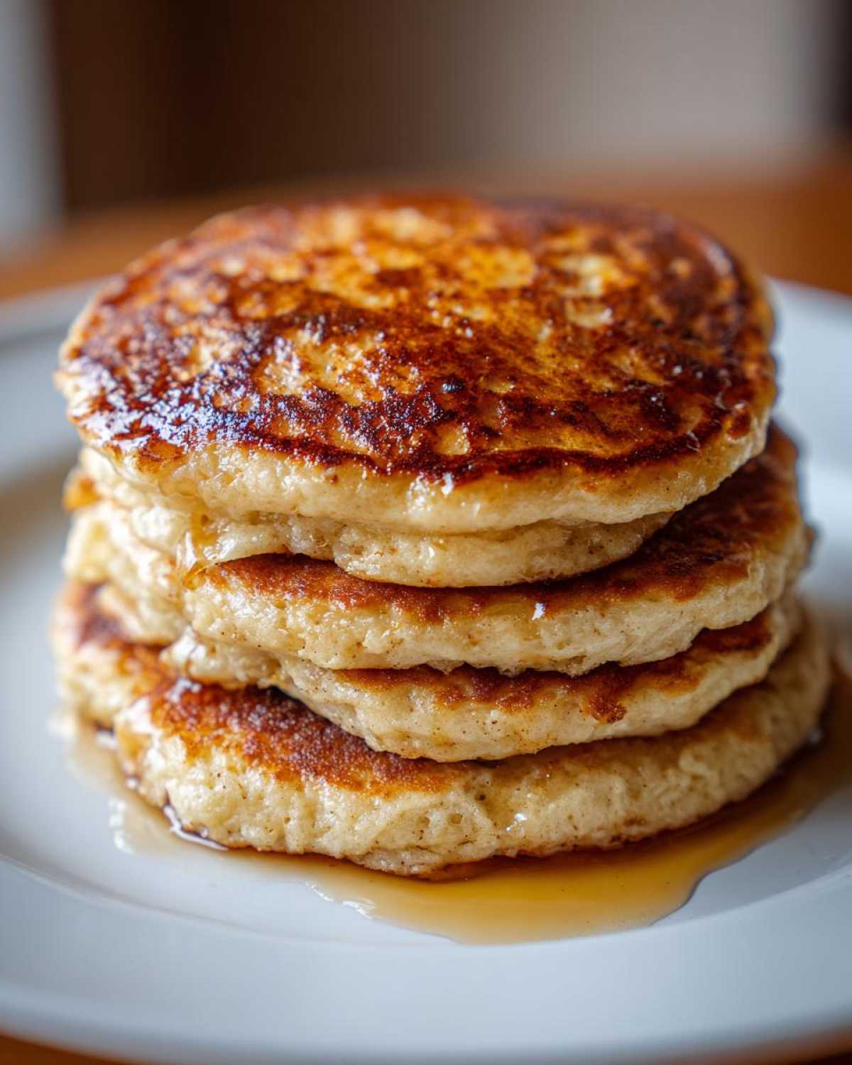 Close-up of a tall stack of golden-brown Vegan Banana Oat Pancakes drizzled with syrup on a white plate.
