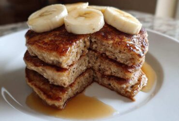 A close-up stack of fluffy Vegan Banana Oat Pancakes drizzled with syrup and topped with fresh banana slices.