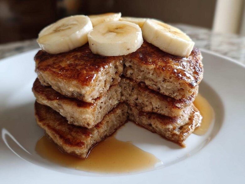 A close-up stack of fluffy Vegan Banana Oat Pancakes drizzled with syrup and topped with fresh banana slices.
