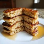 A stack of four thick Vegan Banana Oat Pancakes, cut to show the fluffy interior, drizzled with syrup.