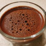 Close-up of rich, dark brown Vegan Chocolate Chia Pudding mixture with small bubbles on top in a clear glass bowl.