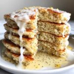 Close-up of a tall stack of fluffy Vegan Lemon Poppy Seed Pancakes drizzled with white glaze and lemon zest.