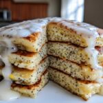 Close-up of a tall stack of fluffy Vegan Lemon Poppy Seed Pancakes cut in half, drizzled heavily with a white lemon glaze.