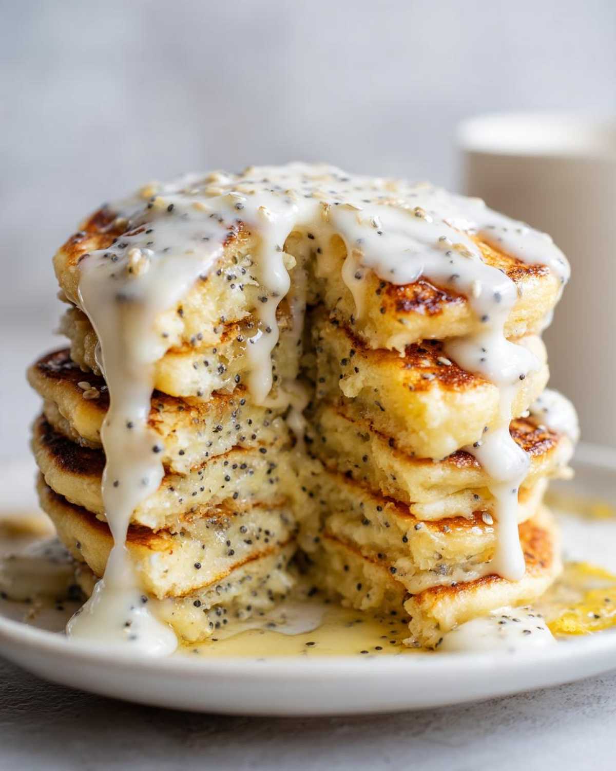 A tall stack of fluffy Vegan Lemon Poppy Seed Pancakes cut open, generously drizzled with a thick lemon glaze and visible poppy seeds.