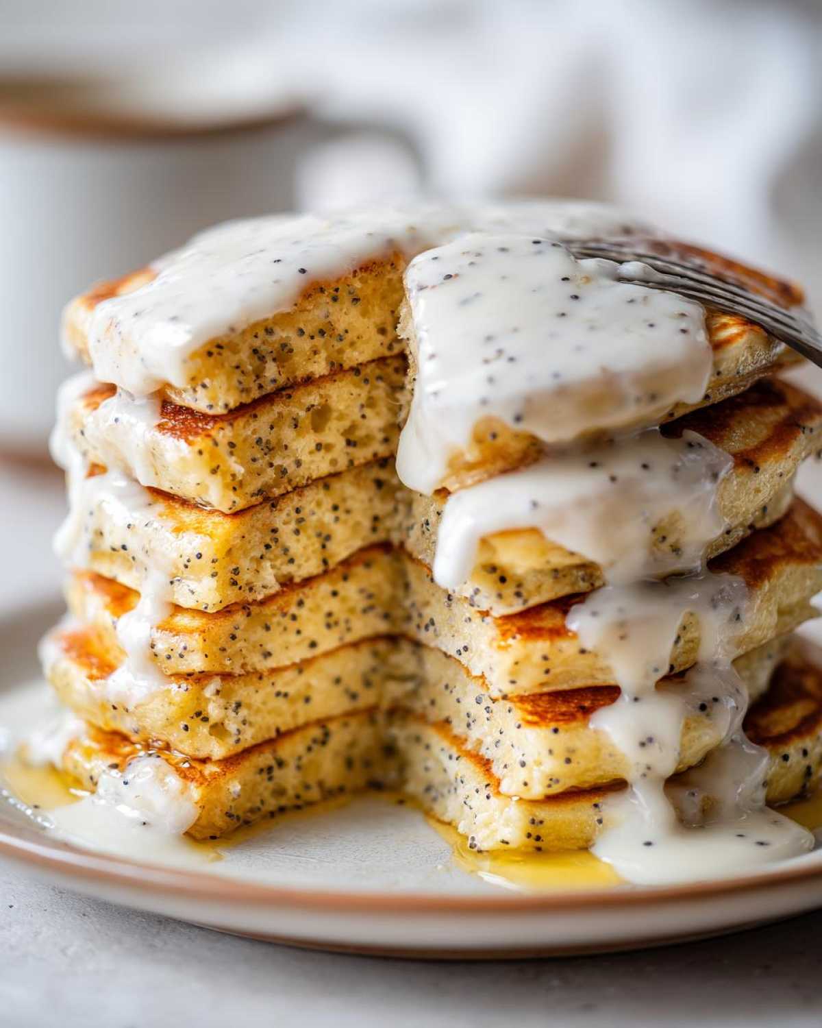A tall stack of fluffy Vegan Lemon Poppy Seed Pancakes covered in a thick, white lemon glaze, with a piece being lifted by a fork.