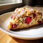 A single, freshly baked White Chocolate Raspberry Scones slice drizzled with white icing on a white plate.