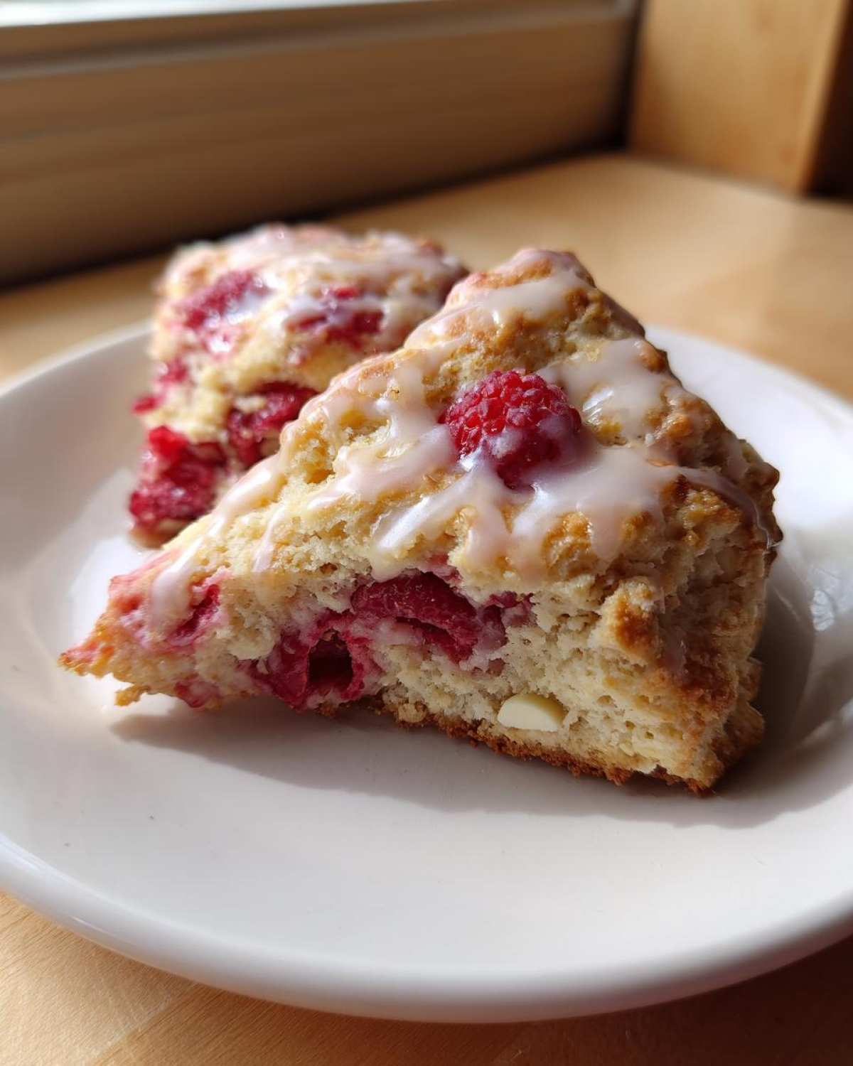 Two triangular White Chocolate Raspberry Scones drizzled with a sweet white glaze, sitting on a white plate.