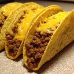 Three hard-shell Cheesy Ground Beef Tacos lined up, filled with seasoned meat and topped with melted cheddar cheese.