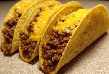 Three hard-shell Cheesy Ground Beef Tacos lined up, filled with seasoned meat and topped with melted cheddar cheese.