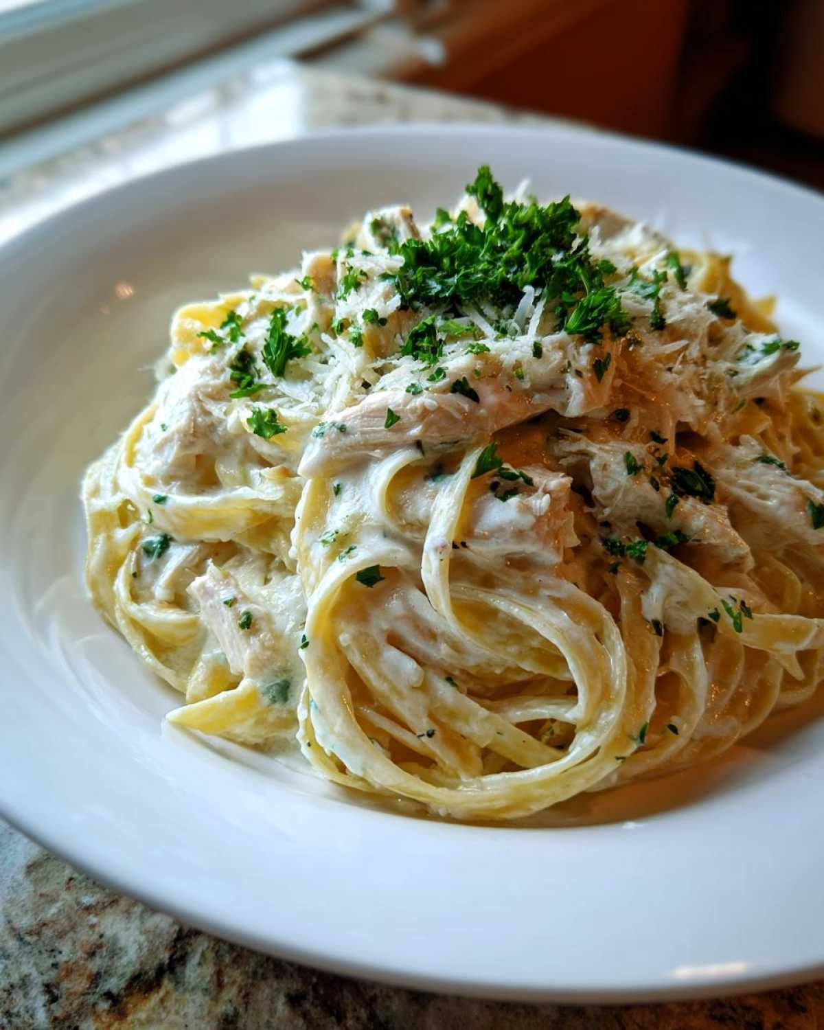 A close-up of a white bowl filled with Creamy Alfredo Chicken Pasta, topped with shredded chicken, Parmesan, and fresh parsley.