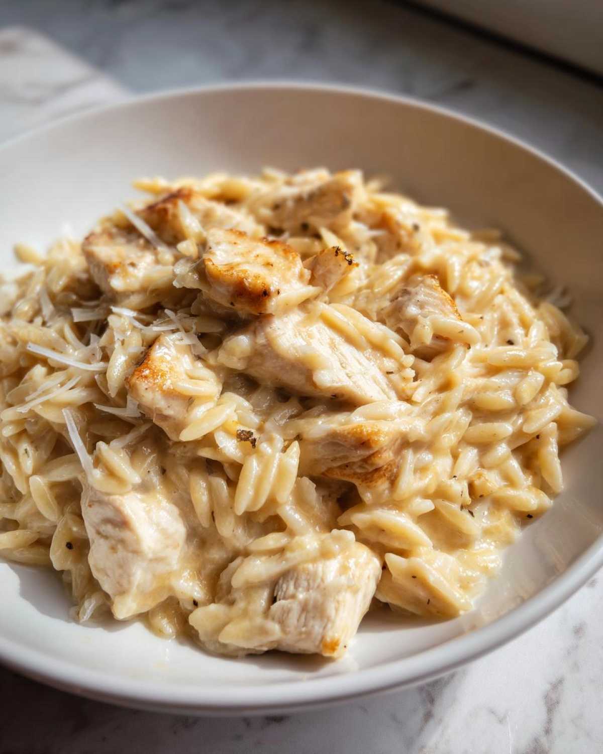 A close-up of Creamy Garlic Parmesan Orzo with Chicken in a white bowl, topped with grated cheese.