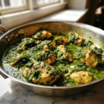 Close-up of chicken pieces simmering in a vibrant green, creamy spinach sauce in a stainless steel skillet.