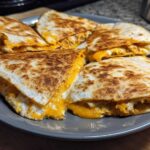 Four wedges of a crispy Cheesy Chicken Quesadilla with melted orange and white cheese spilling out, served on a grey plate.