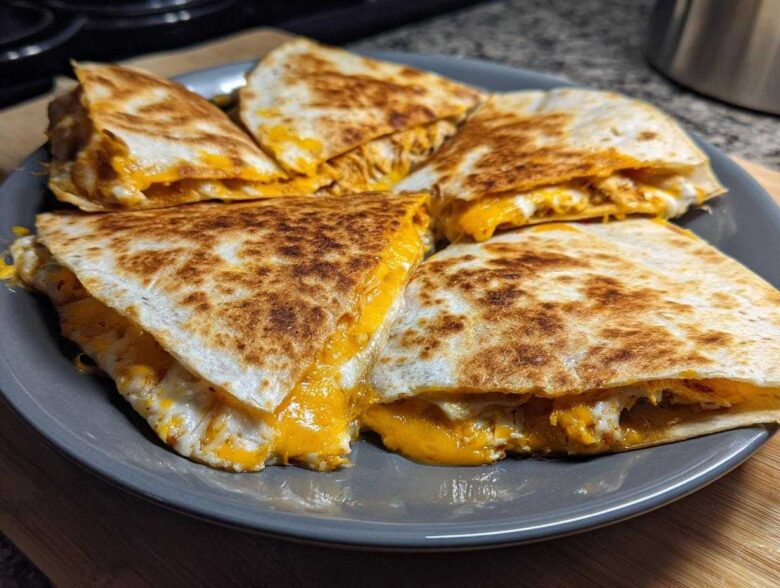 Four wedges of a crispy Cheesy Chicken Quesadilla with melted orange and white cheese spilling out, served on a grey plate.