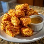 A pile of golden, crispy chicken nuggets served on a white plate next to a small ramekin of honey garlic dip.