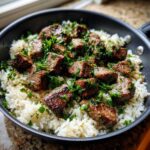 Close-up of juicy steak bites served over white rice, topped with fresh parsley in a black skillet, ready for Garlic Butter Steak and Rice Skillet Dinner.