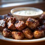 A plate piled high with glistening Garlic Butter Steak Bites served alongside a small bowl of creamy garlic sauce.