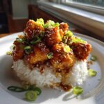 A serving of sticky Honey Garlic Chicken Bites piled high on a mound of white rice, topped with sesame seeds and green onions.