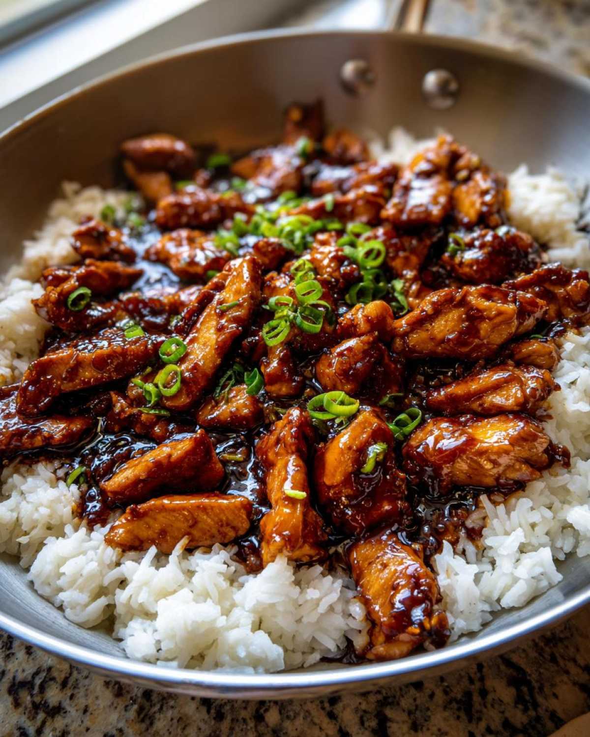 Glazed pieces of chicken coated in dark honey garlic sauce served over white rice in a skillet, garnished with green onions.