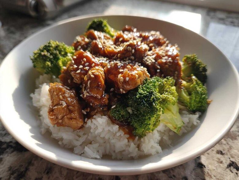 A close-up of a Honey Soy Chicken Bowl Quick Dinner Idea featuring glazed chicken pieces over white rice and bright green broccoli florets, topped with sesame seeds.