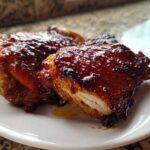 Close-up of two glistening Honey Soy Chicken Thighs with crispy, caramelized edges on a white plate.