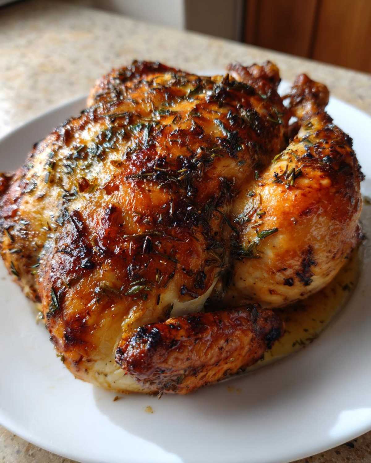 A whole, perfectly roasted chicken with dark golden, crispy skin seasoned generously with herbs, resting on a white plate.