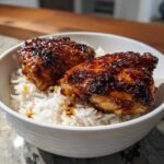 Two glazed Sticky Sweet Chili Chicken Thighs resting on a bed of white rice in a white bowl.