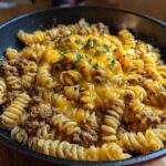 Close-up of Cheesy Ground Beef Pasta Skillet with melted cheddar cheese and parsley garnish in a dark skillet.