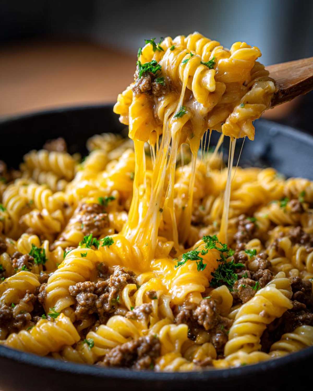 A wooden spoon lifts a portion of Cheesy Ground Beef Pasta Skillet, showing long, melted cheese strings stretching back into the pan.