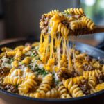 A wooden spoon lifts a portion of Cheesy Ground Beef Pasta Skillet, showing long, melted cheese strings stretching from the pan.