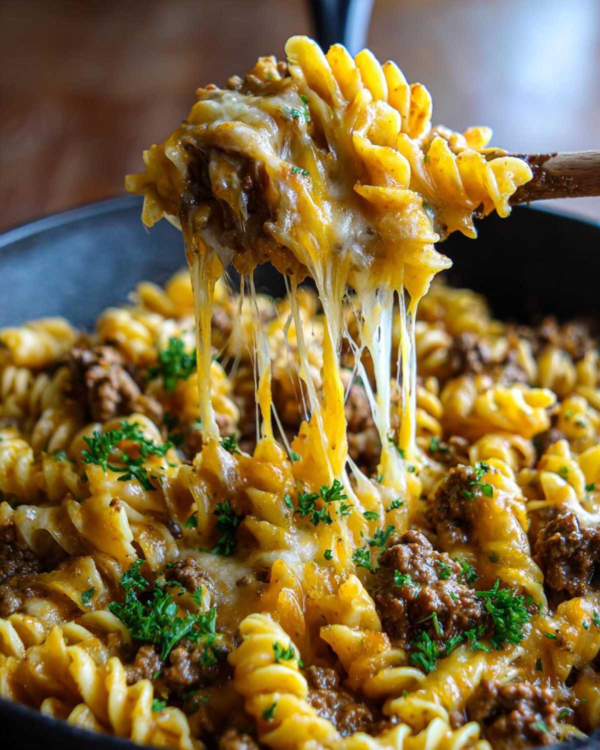 A spoonful of Cheesy Ground Beef Pasta Skillet shows a dramatic cheese pull over the skillet of pasta, beef, and garlic sauce.