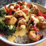 Close-up of a Honey Garlic Chicken Bowl featuring glazed chicken pieces, broccoli, red peppers, and a generous drizzle of creamy sauce over rice.