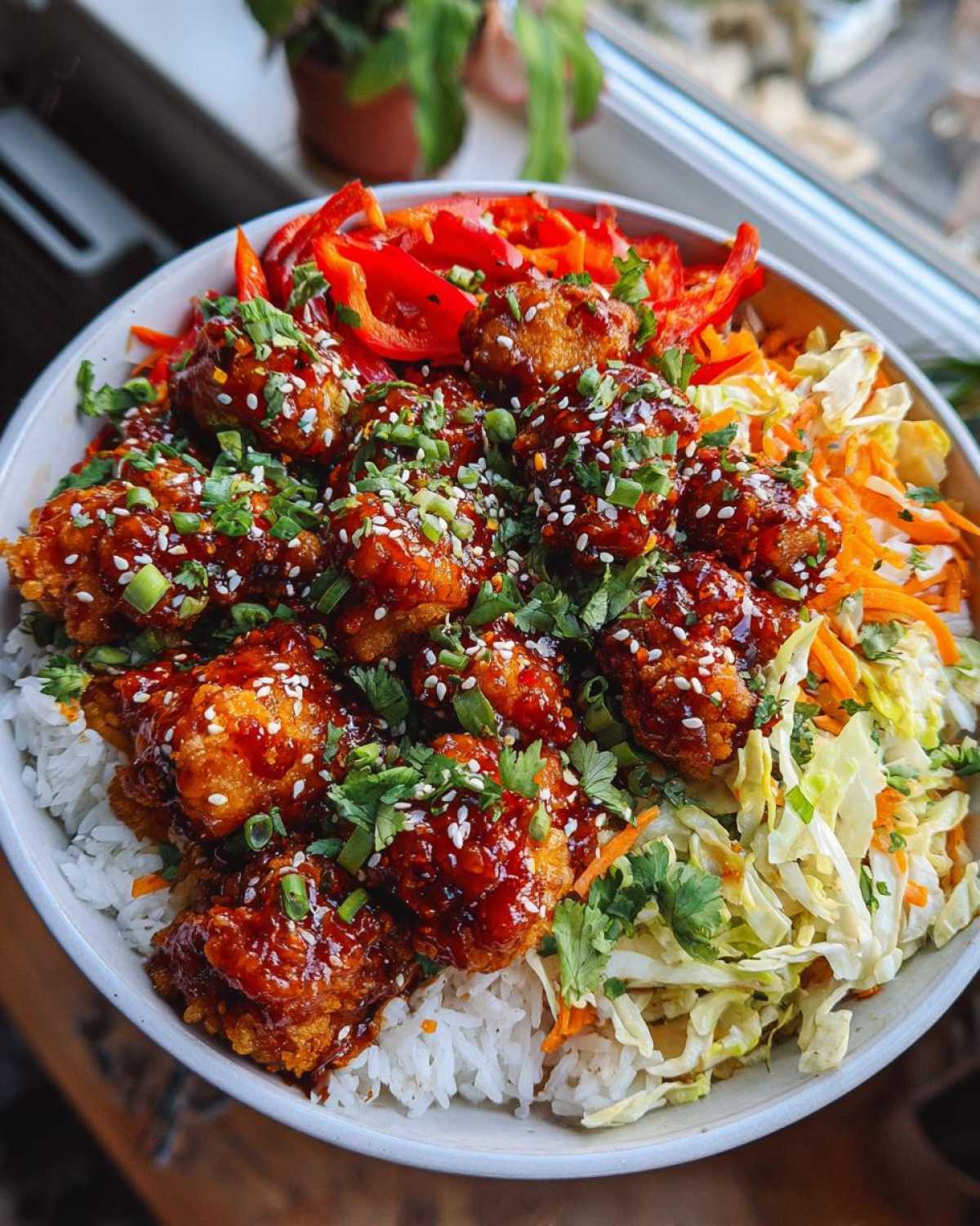 A close-up of a Sweet Chili Chicken Bowl featuring glazed chicken pieces over rice, topped with sesame seeds, green onions, and crunchy slaw.